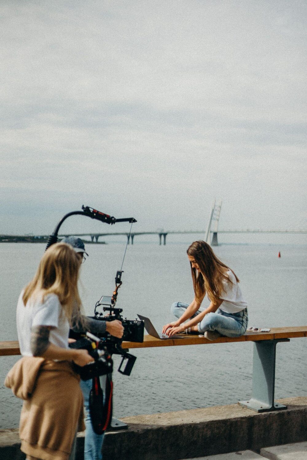 A film crew filming an interview outdoors by a waterfront, one woman holding a camera and another seated on a railing