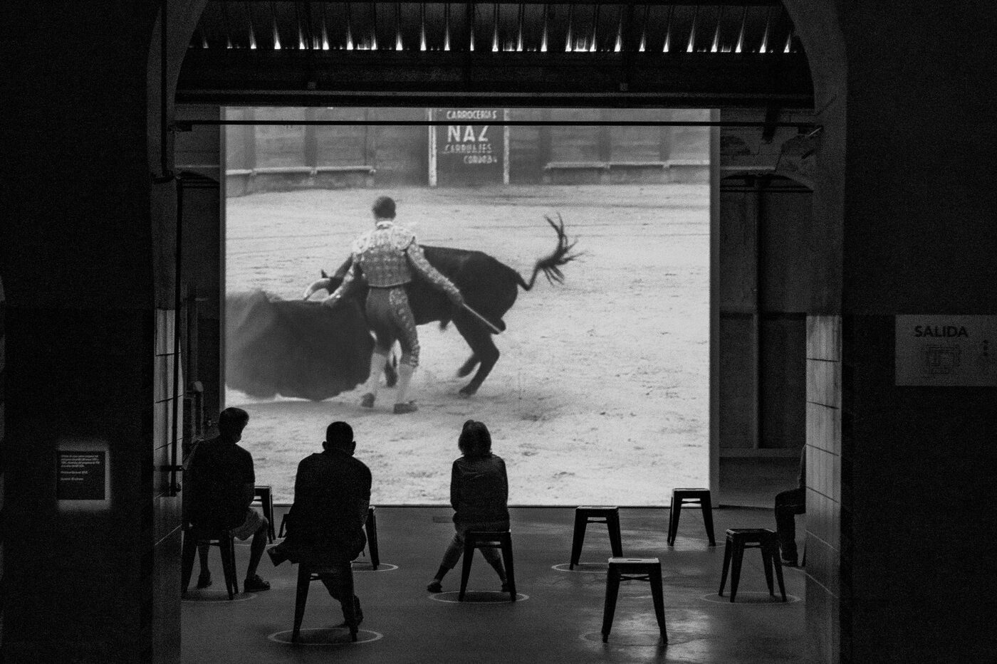 Silhouettes of people watching a bullfighting film screening from stools inside an old theatre archway